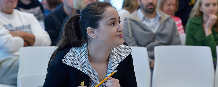 A female student dressed in business fashion, in a seated crowd of students, looks and listens intently. She is holding a pencil.