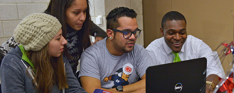 Four students of diversity sit in front of a laptop and together look at the contents on the screen. One of male students points with a smile at something unseen on the screen.