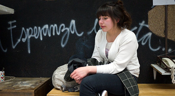 A female student sitting in rest, holding her jacket in her lap with her backpack at her side. A black wall behind her reads, "Esperanza", which more text behind her. There is a white telephone on the right, and a coffee cup with hearts on it to the left.