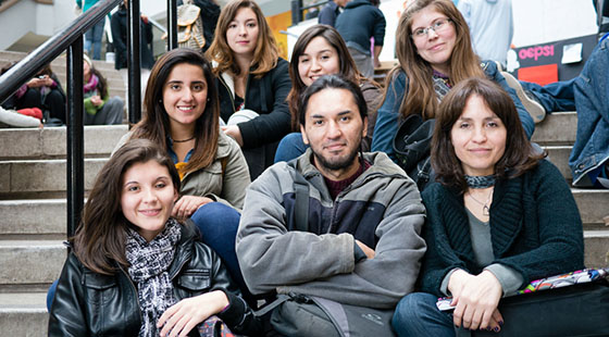 A group of students of diversity sitting on ascending concrete stair steps with a teacher. They are posed with smiles and dressed for cold weather.