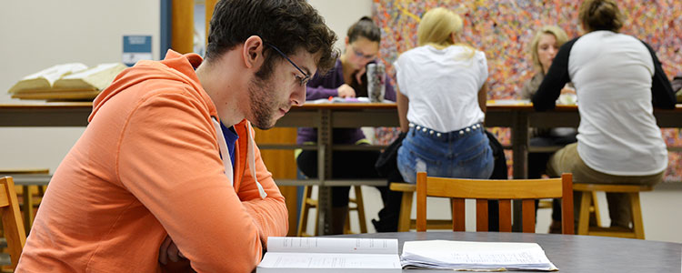 A male student wearing a bright orange hoodie looks intently at a text book in a colorful study area. Four female students sit at another table in the background, slightly blurred due to perspective, who are studying together.