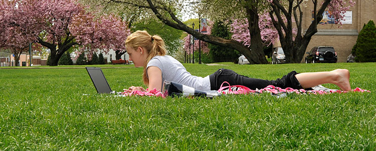 A female student lies on a towel on grass, doing school work on her laptop. Pink and green trees fill the background, as well as a visual of WCSU's White Hall and some parked cars.