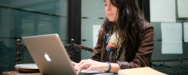 A female student, with long dark hair and wearing a brown coat and dull colored scarf, sits typing on her Mac laptop.