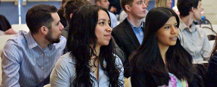 A crowd of diverse students, dressed in business casual fashion, sit in rows of chairs and watch and listen intently to a special speaker.