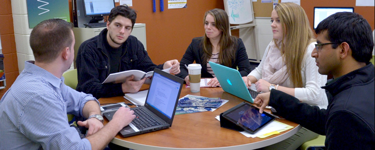 Five students of diversity sit at a study table, with laptops, tablets, and textbooks in hand, studying for an upcoming assignment or project.