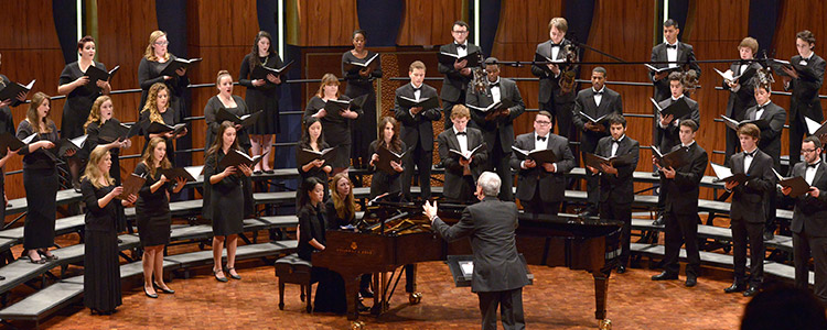 A choir, made up of students wearing tuxedos and black dresses, sings in one of WCSU's gorgeous amphitheaters. Two female student musicians sit at a grand piano in front of the conductor.