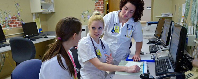 Three female nursing students chat in front of a textbook in one of the nursing offices. They sit at a desk topped with rows of computers and telephones.