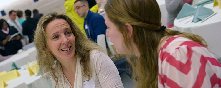 Two female students converse at a dinner event, where tables are covered in white table clothes and blue folded napkins. Other students of diversity can be seen in the distance chatting or setting up for the event.