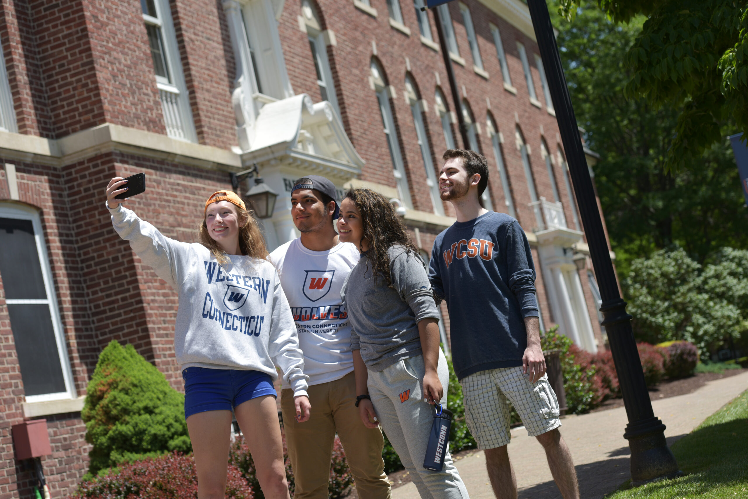 Four students taking a selfie in front of a campus building. All students are wearing Western CT state clothing.