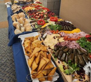 Catered buffet table featuring breads, cheeses, meats, fresh fruit, and vegetables, with event decorations in the background.