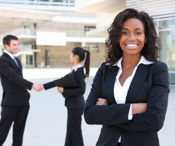 Lady in business attire smiling and crossing her arms in a confident pose as two other business people in the background shake hands