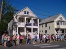 A large group of people are lined up along a street in front of two houses. Trees can be seen on the right and across walk lines the street.