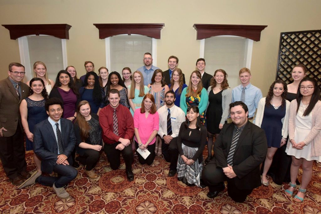 Many students in formal attire stand together to take a photo. There are three windows with blinds down behind them. They are standing on a red carpet with an intricate circular pattern.