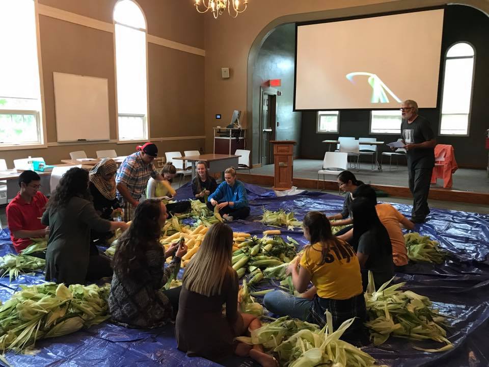 Thirteen students shucking corn on a large purple tarp covering the Honors Lyceum floor. The professor stands at the front of the room with a paper in hand.