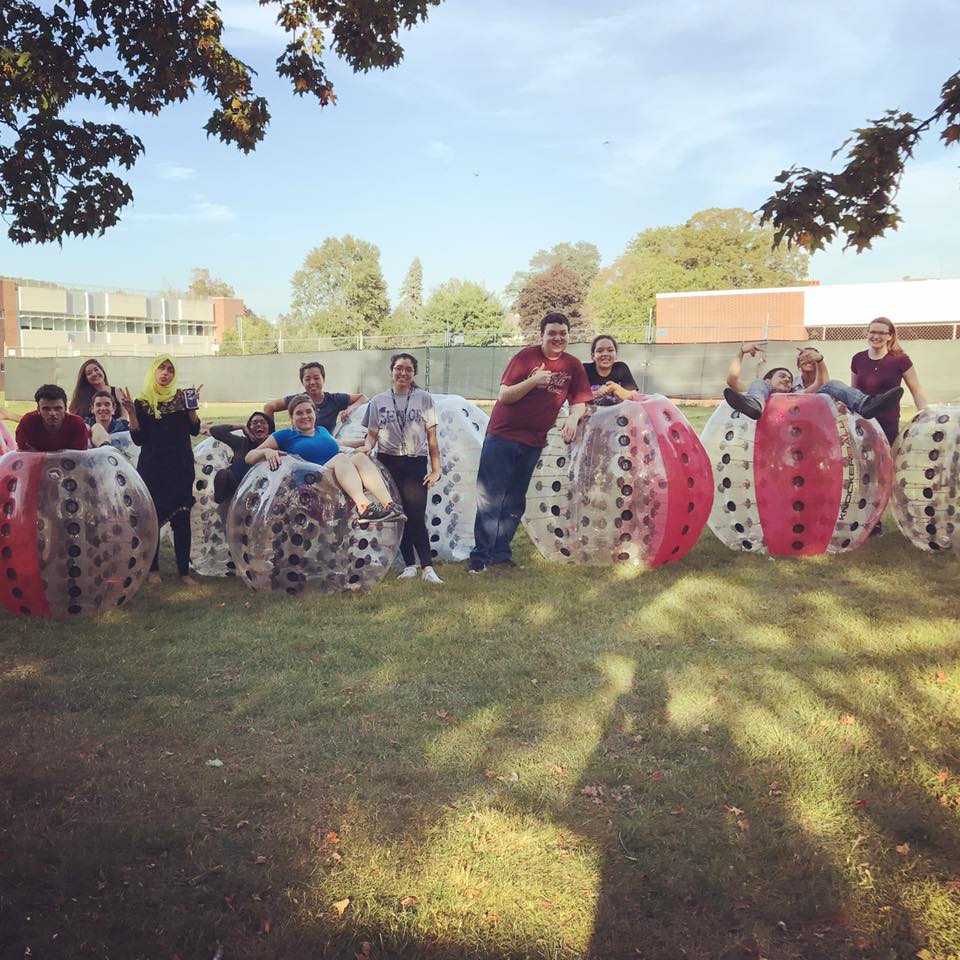 Students standing in and leaning on large plastic bubbles for bubble soccer on the lawn between the Honors House and the Science Building. The bubbles are half red and half clear.