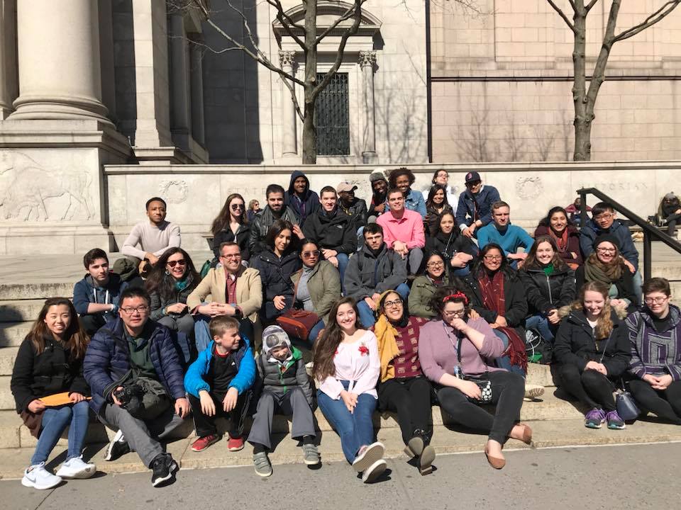 Many students sitting on front steps of American Museum of Natural History for a group photo. They are all wearing jackets and sweatshirts.