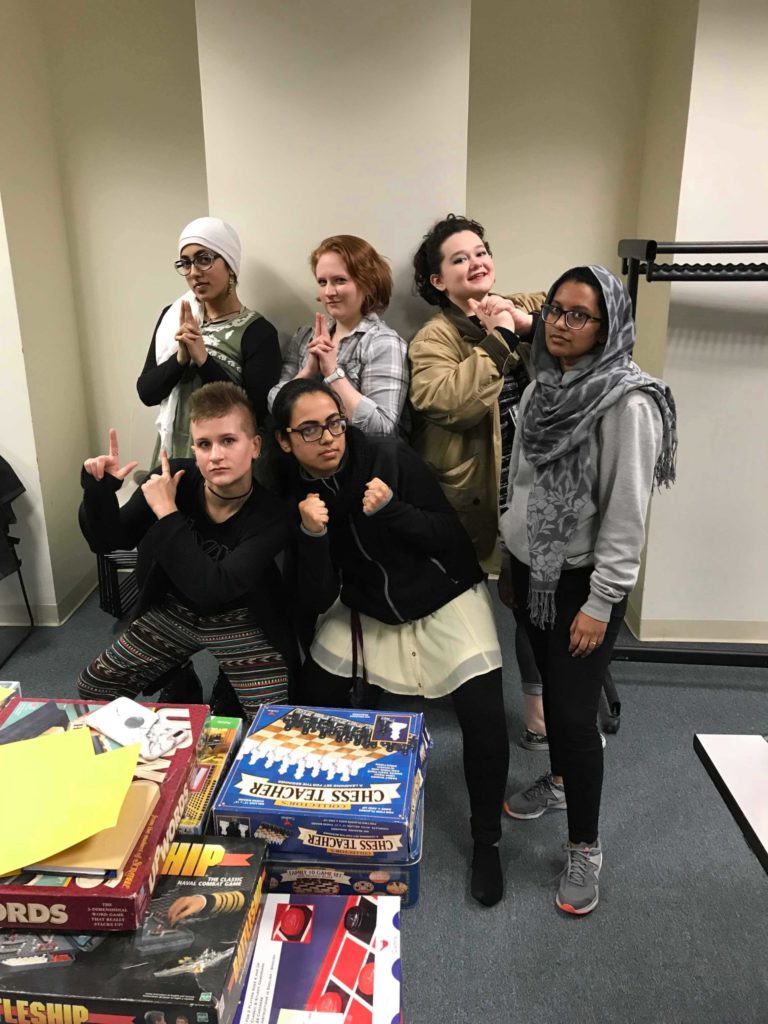 Six students standing together and striking very cool poses in a group photo. There are various board games on a table in front of them.