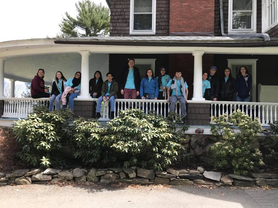 Students standing on and sitting on railing of wrap-around porch of building for a group photo. There are a few bushes with very leafy branches in front of them.