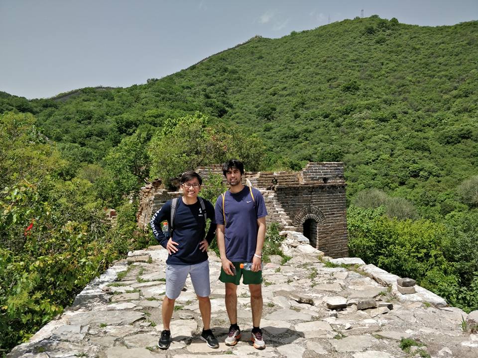 Hieu is on the left, and his friend is on the right. They are standing in the middle of a stone path on the Great Wall of China. There is a giant mountain behind them with many trees.