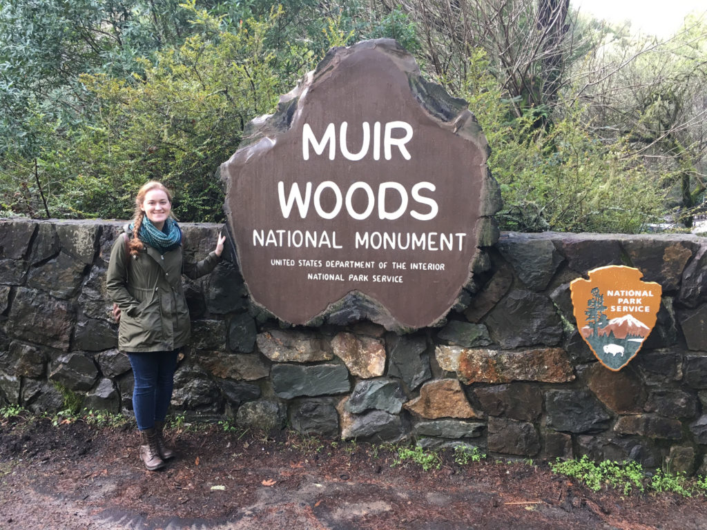 Shannon stands next to a large faux slice of tree stump on its side. "Muir Woods National Monument, United States Department of the Interior, National Park Service" is written in capitalized white font on the inner bark. The National Park Service seal is on the stone wall on the right. Shannon is wearing a blue infinity scarf, an olive green jacket, blue jeans, and brown boots.