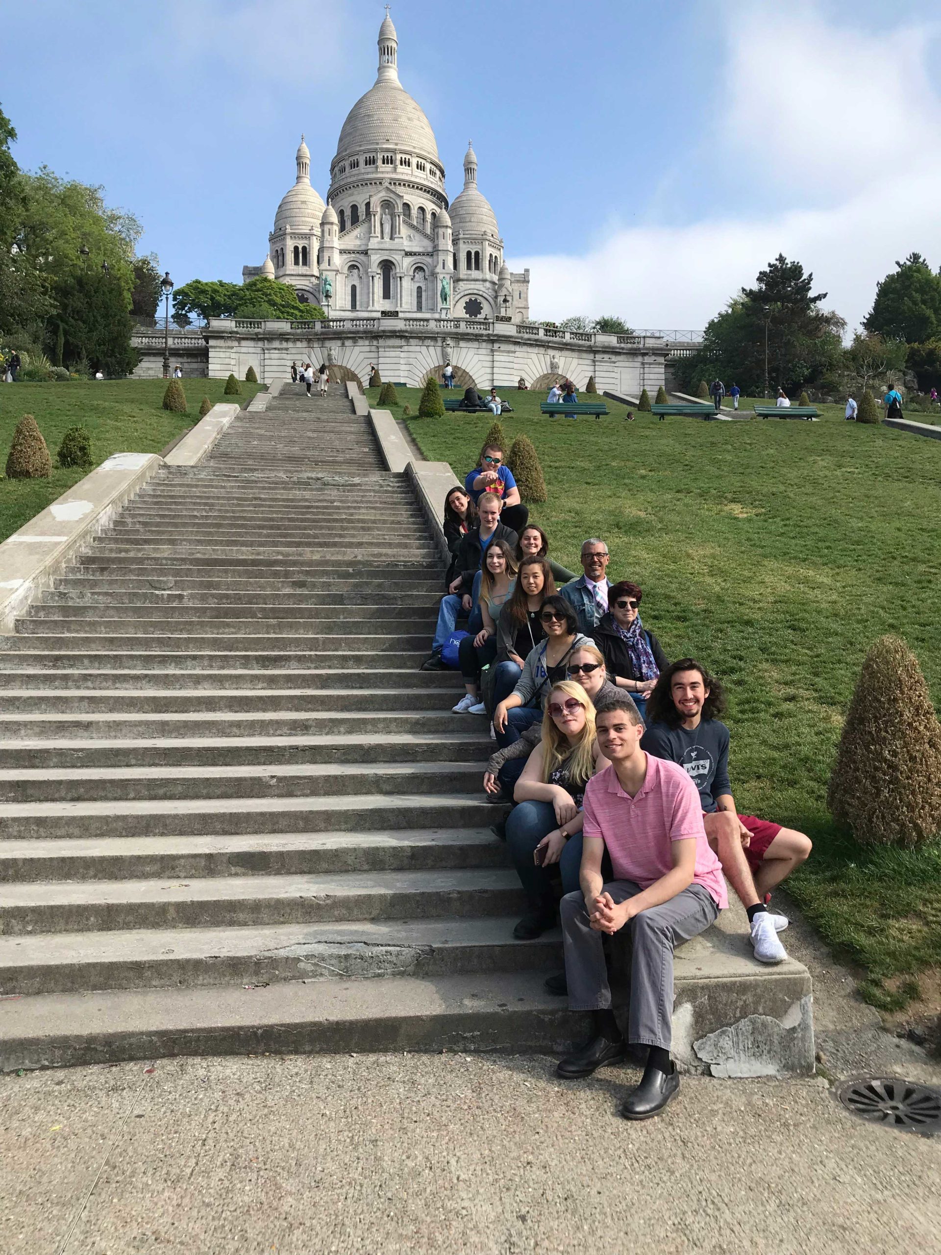 Students and professors sit on the bottom of the wide concrete rail on the right of the steps. The Sacre Coeur Basilica is at the top of the steps, and there is a lot of greenery in the park.