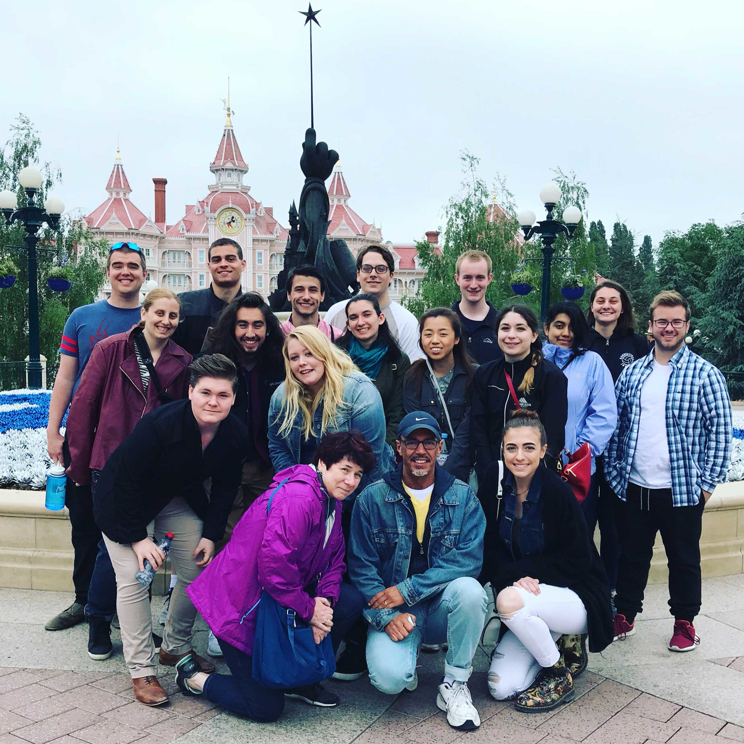 Students and professors taking a group photo in front of a castle at Disneyland Paris. There are a few clusters of blue and white flowers behind them.