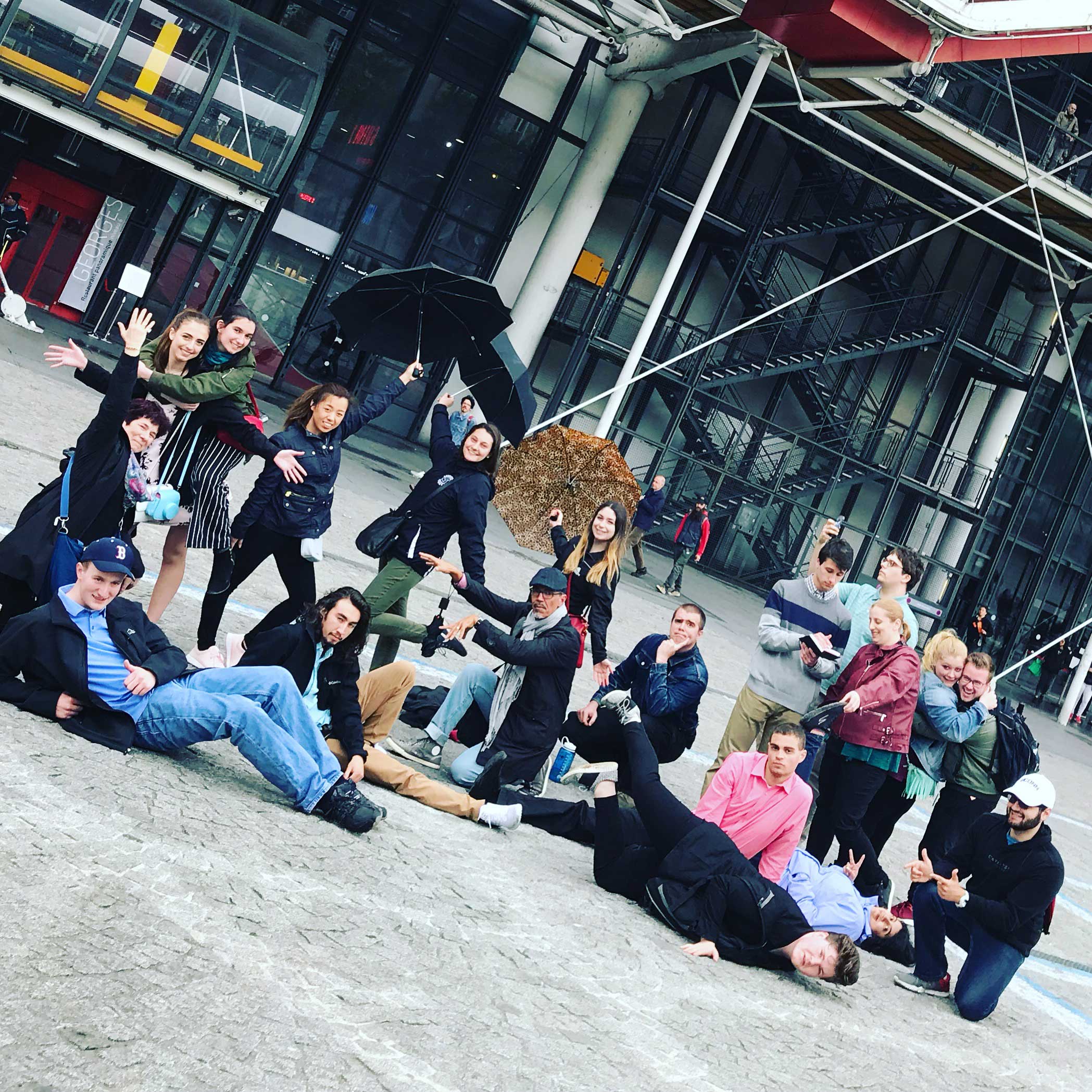 Students and professors pose outside a large entertainment building. They're either lying on the ground, hugging each other, holding up umbrellas, and striking other very cool poses.