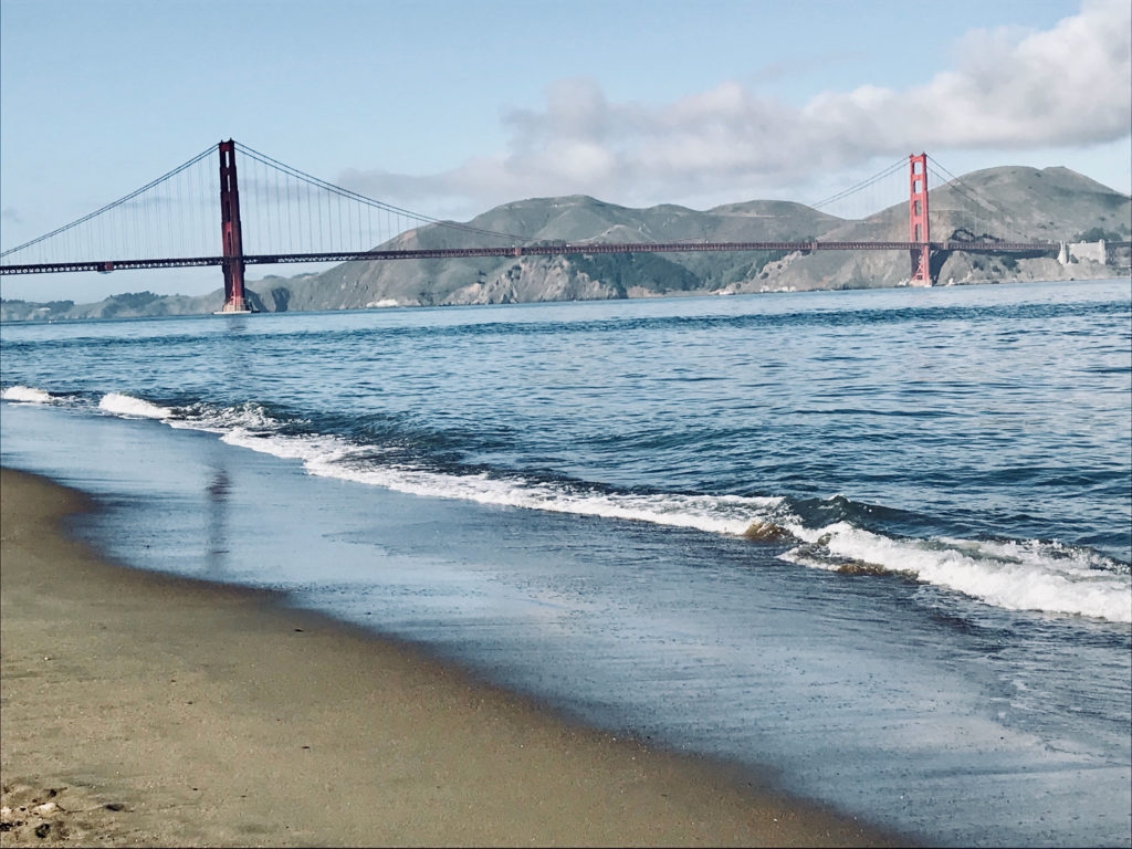 The tide recedes back into the water. The golden Gate Bridge is off in the distance, and there are a few small mountains behind it.