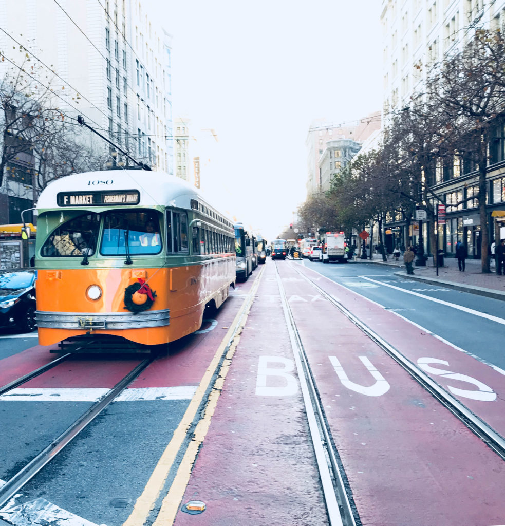 Taken from the middle of a street. A white and yellow street car is to the photographer's left, and there is empty road on the right. The road is lined with leafless trees on either side.