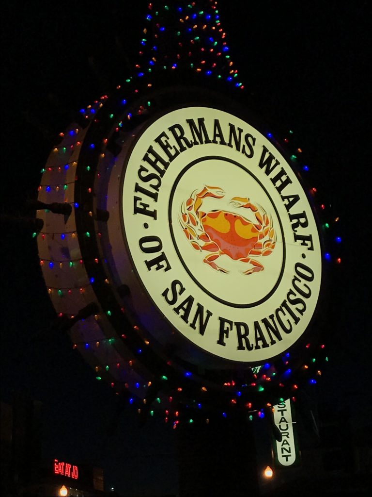 Fisherman's Wharf of San Francisco is written around the circular sign, and a yellowish-orange crab is in the center. There are many tiny dot lights of various colors around the sign as well.