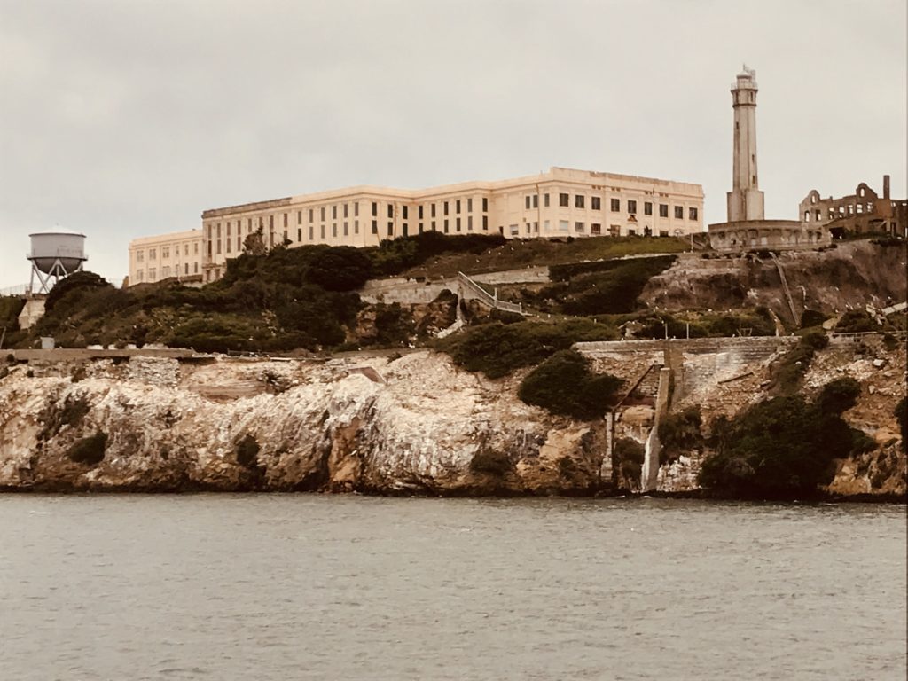 Alcatraz, a large, light tan building, sits at the top of a large rocky island. A water tower sits on the left, and a large white watchtower sits on the right. There is a short wall around the island border, and there are many large white rocks at the base.