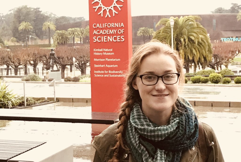 Shannon stands in front of a tall, rectangular pillar that says, "California Academy of Sciences," capitalized in white font. Shannon is wear a dark green infinity scarf, glasses, and an olive green jacket.