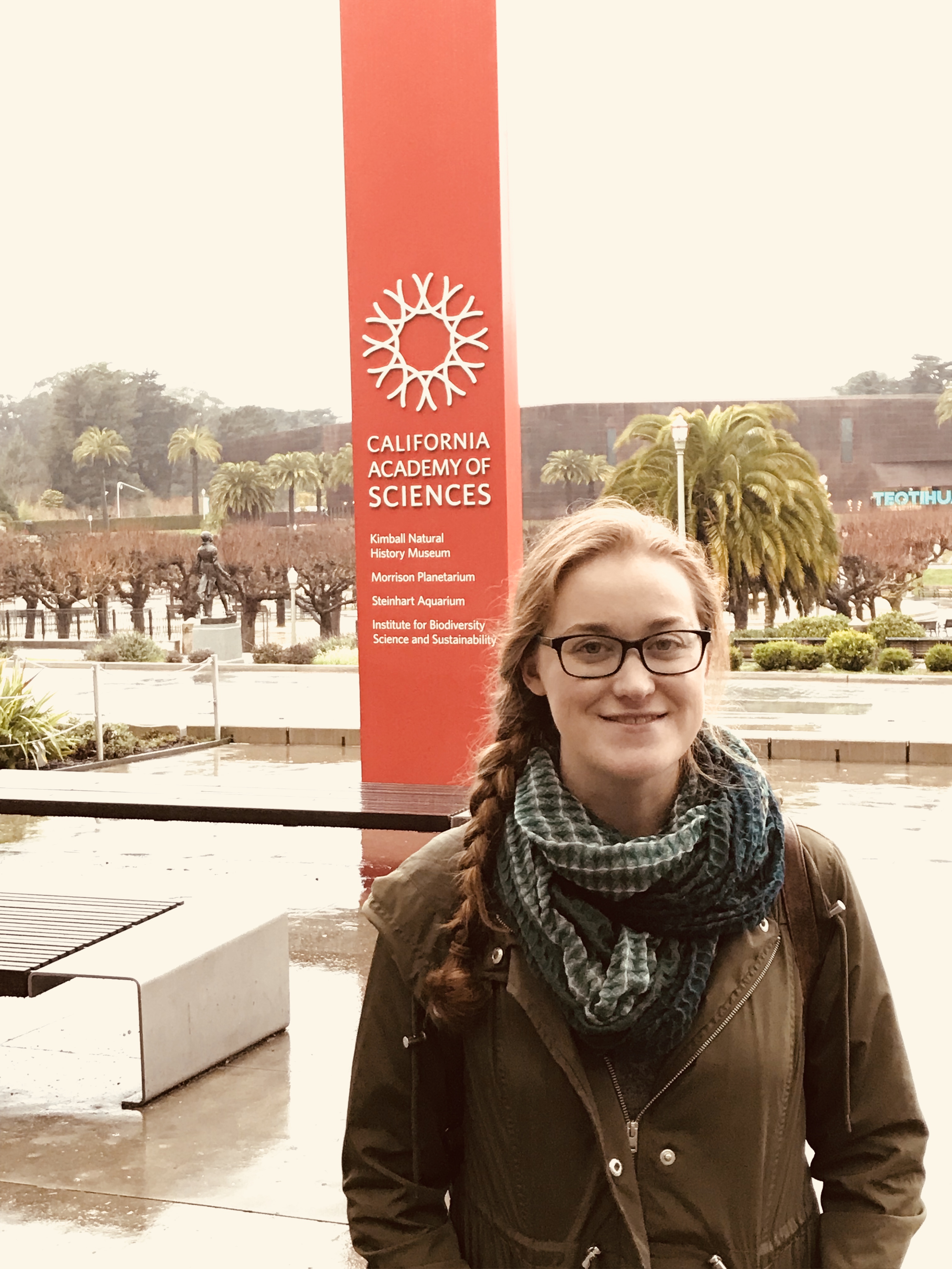 Shannon stands in front of a tall, rectangular pillar that says, "California Academy of Sciences," capitalized in white font. Shannon is wear a dark green infinity scarf, glasses, and an olive green jacket.