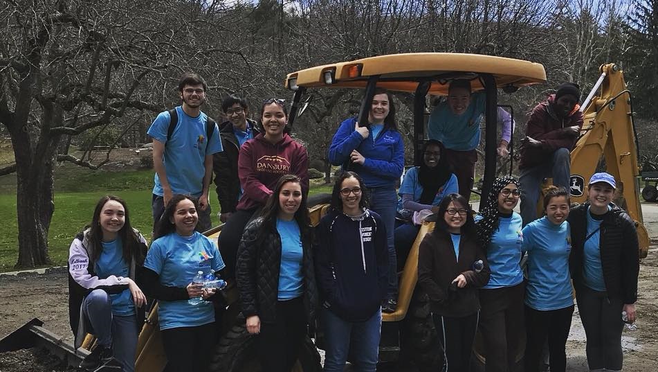 Students stand near and on a construction vehicle to take a photo. They are all wearing light blue shirts with the multi-color Day of Service logo on it.