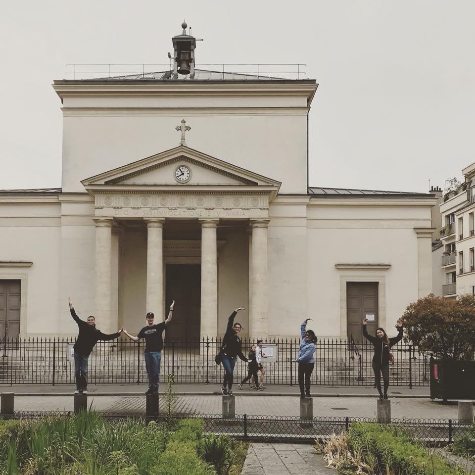 Students standing in front of Sainte-Marie des Batignolles Church. They're holding their arms up in the air kind of like they're doing the YMCA. The church is a large white building with a small clock and cross above the main entrance.
