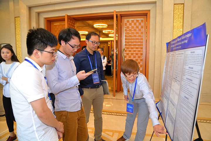 A presenter shares their research on a poster to three audience members. Everyone looking at the poster is wearing glasses. They are wearing dress shirts and pants.