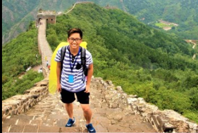 Hieu is wearing a blue and white striped shirt, blue shorts, blue sneakers, glasses, and a backpack. He is standing on steps going up the path of the Great Wall of China. You can see more of the Great Wall behind him, winding over and around mountains.