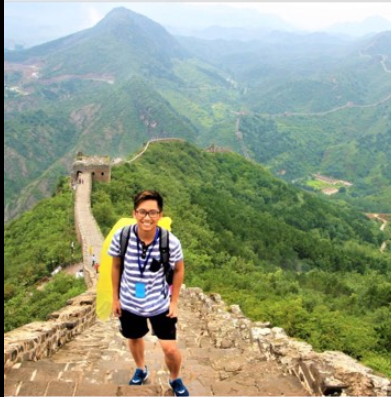 Hieu is wearing a blue and white striped shirt, blue shorts, blue sneakers, glasses, and a backpack. He is standing on steps going up the path of the Great Wall of China. You can see more of the Great Wall behind him, winding over and around mountains.