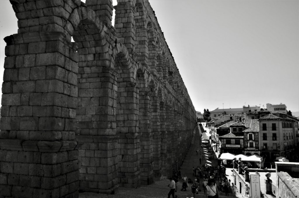 The aqueduct is made up of many large stone bricks that create arches. There are many smaller buildings on the right side, and people walk up the steps in the middle.