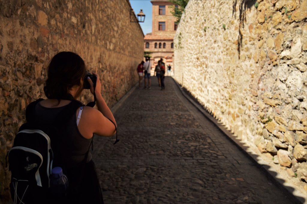 Rachel stands between two walls made of stone on a stone path. There are people standing towards the end of the path. Rachel has a backpack on her back with a plastic bottle in the right outer pocket.