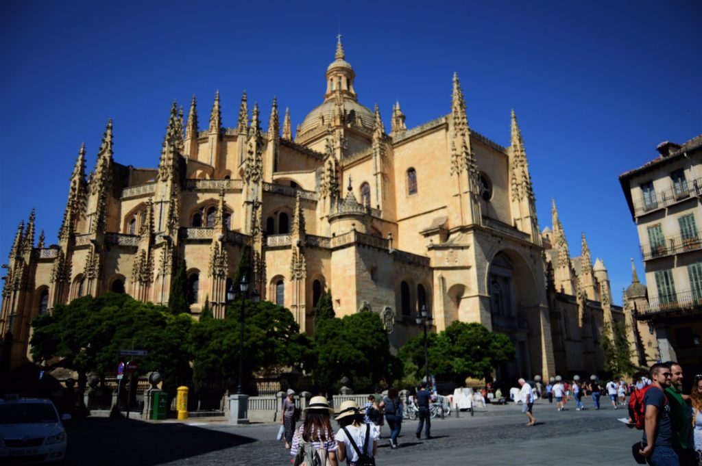 The Segovia Cathedral is a light tan church with many spiked turrets and windows into many rooms. There are a few very green short (by comparison) trees in the front, and people walk around the plaza outside.