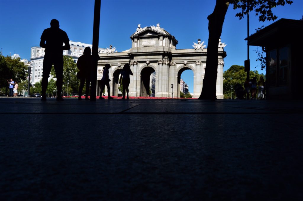 The Puerta de Alcalá is a gray monument in the background. It is made of many gray stone blocks. There are many carvings on the top that look like wings popping out of circles from far away. There are two circles above each of the three arches and a light above each circle.