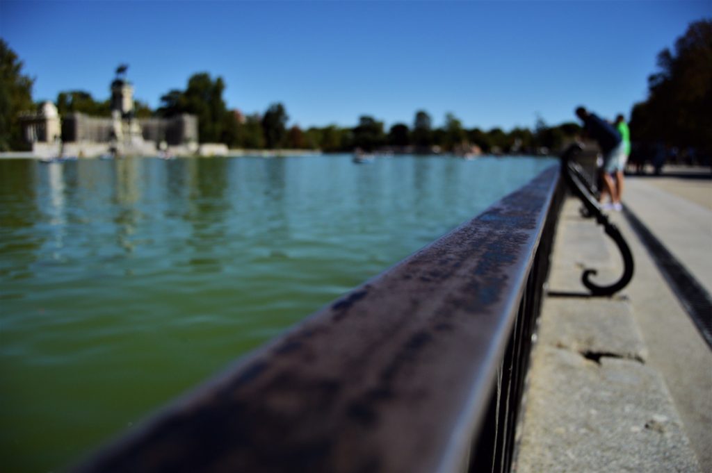 There is a somewhat rusty railing around the lake in the foreground. Everything else is blurred, which includes the monument in the background, the lake itself, and the many trees around the lake.