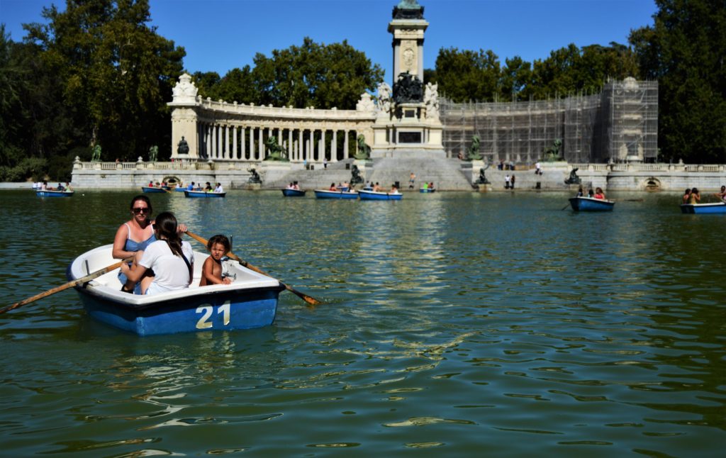 The El Retiro monument is in the background. El Retiro is a large half circle made of many covered pillars surrounding an obelisk-like structure in the middle. The right side of the pillar arch has scaffolding around it. There are also many blue rowboats with people in them on the lake.