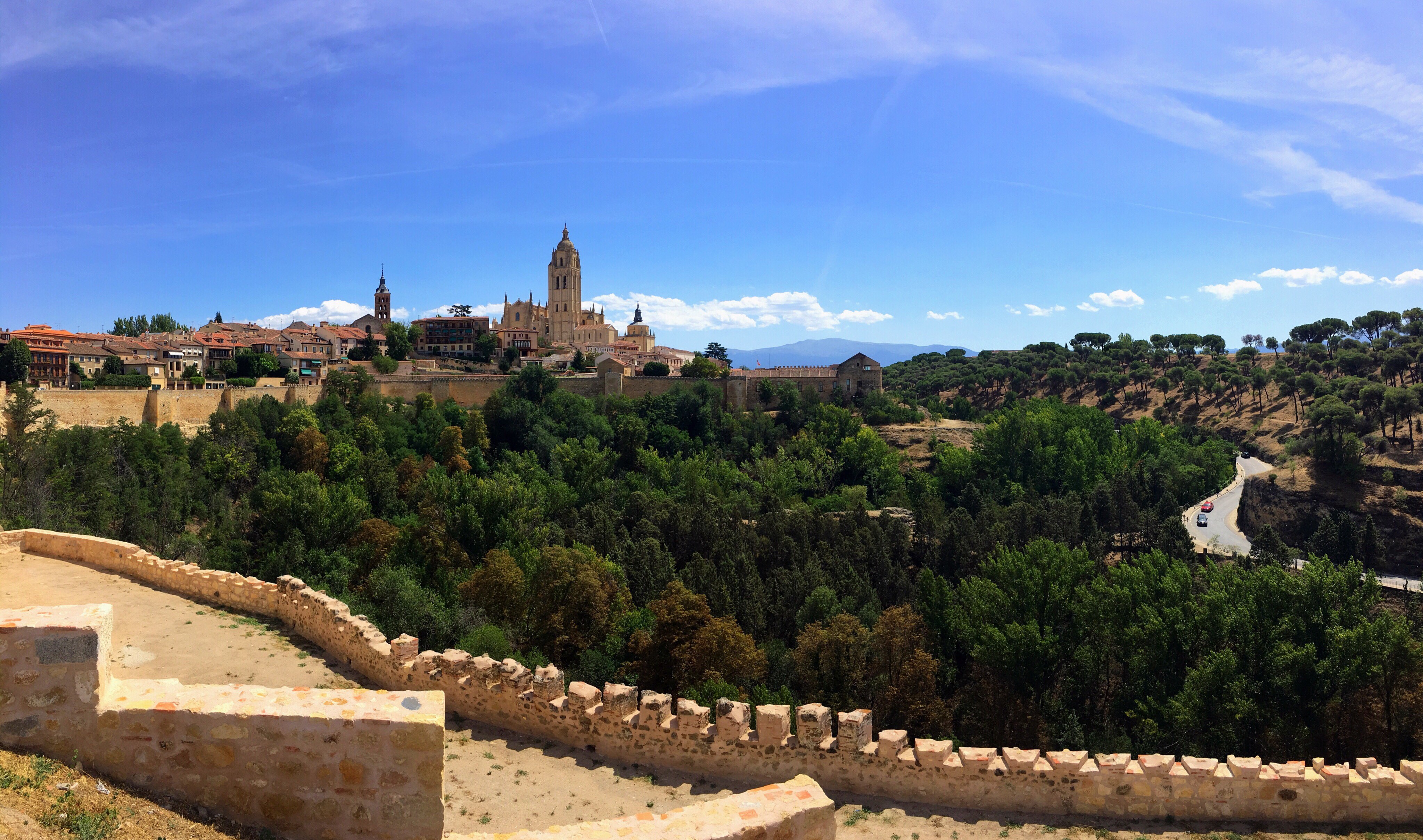The photographer is standing near a stone wall across from the fortress (or castle) of Segovia. There are many trees in the divide between the wall and the fortress.