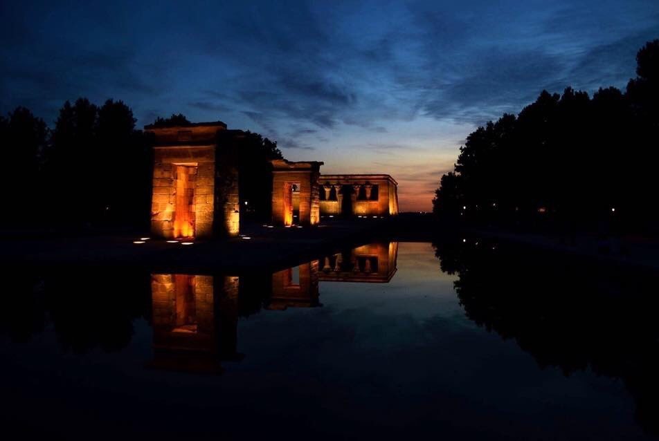 The Temple of Debod has lights on the ground that illuminate the walls of the temple. The sun is disappearing under the horizon, and the sky is mostly dark blue. There is water around the temple and trees around the water.