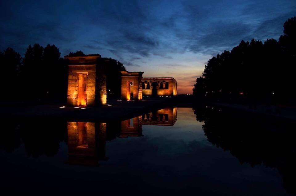 The Temple of Debod has lights on the ground that illuminate the walls of the temple. The sun is disappearing under the horizon, and the sky is mostly dark blue. There is water around the temple and trees around the water.