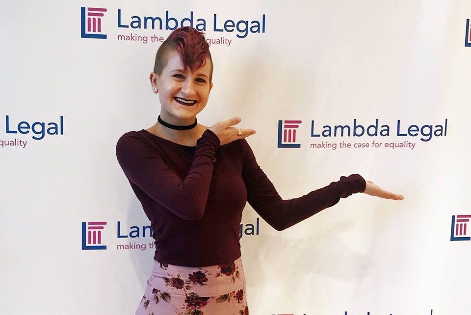 Erika is standing in front of a backdrop with the Lambda Legal logo on it (a blue vertical and horizontal line forming a right angle with a pink pillar inside) and Lambda Legal's tagline: "making the case for equality." Erika is wearing a long sleeve maroon shirt and a pink skirt with roses on it over black dress pants. She is smiling and gesturing her hands around the Lambda Legal logo.
