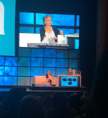 Francoise Mouley sitting on a couch on a stage while speaking to another presenter at the conference. A video of her speaking is projected on a screen above the stage with captions.