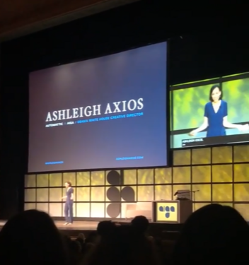 Ashleigh stands in the middle of the stage with the title slide of a Powerpoint containing her name projected on a screen. A live video feed of her is on a screen above the stage with captions. She is wearing a blue short sleeve shirt and jeans.